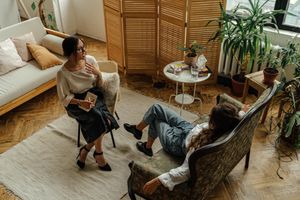 Woman in white long sleeve shirt sitting on brown wooden armchair in a coaching session with another woman sitting on a sofa